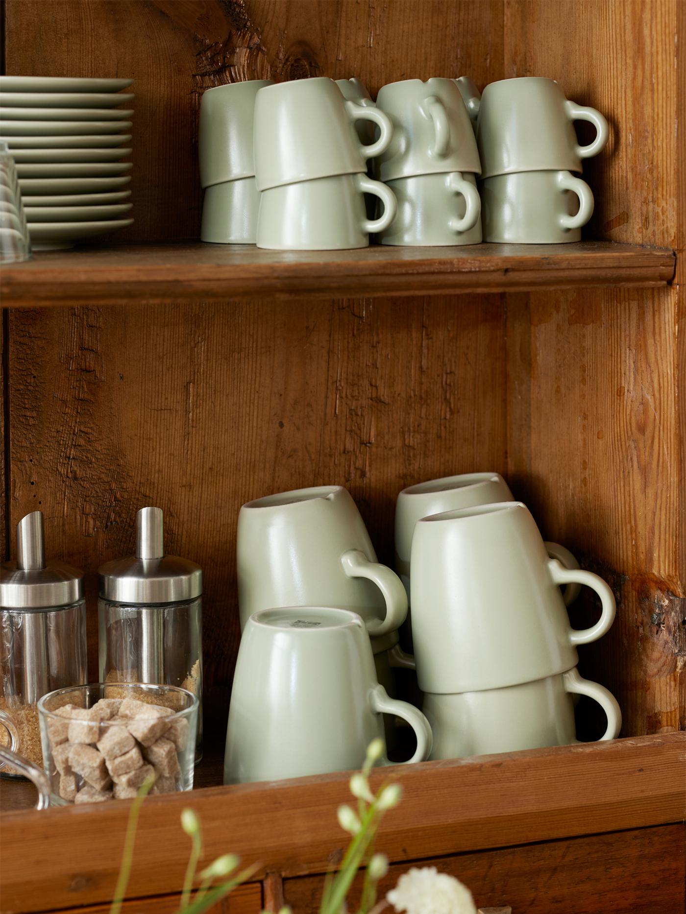 Matt green FÄRGKLAR cups, mugs and saucers and two DOLD portion sugar shakers are stored on two shelves in a cabinet.