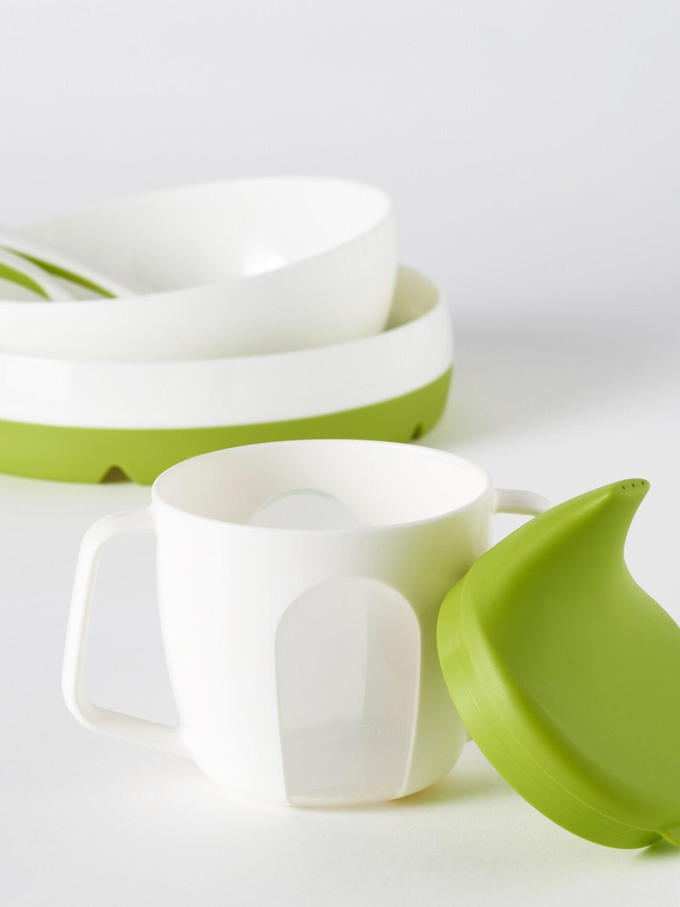 เด็กผู้ชายสวมผ้ากันเปื้อน MATVRÅ/มาทรัว นั่งบนเก้าอี้สูงที่โต๊ะซึ่งมีจาน แก้วมัค และช้อนส้อมมีดวางอยู่