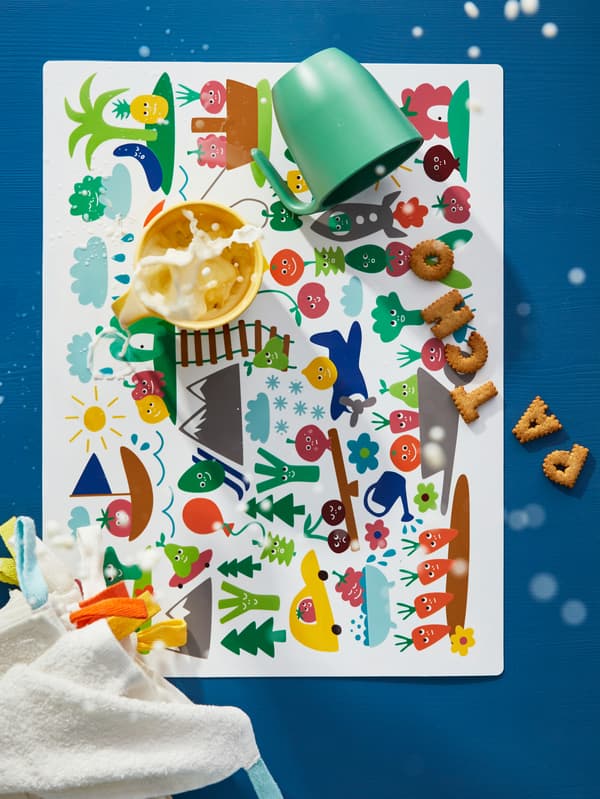 MATVRÅ kids place mat with colourful vegetable figures and children’s plastic beakers with flying milk, biscuits and cloths.