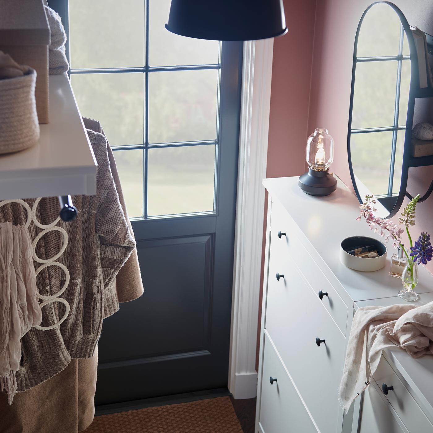 مدخل يحتوي على خزانتين للأحذية بلون أبيض عليهما منتجات متعددة، بالإضافة إلى علّاقة قبعة بلون أبيض ومرآة سوداء مع وحدة تخزين.