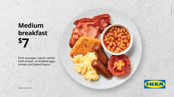 Medium breakfast. White plate on concrete background with scrambled egg, 2 sausages, 1 hash brown, bacon, fried tomato, and small bowl of baked beans, for $7
