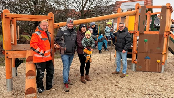 Mehrere Erwachsene und Kinder stehen und sitzen an einem neuen Holzspielgerät auf einem Spielplatz. Zu sehen sind Kletter- und Balancerelemente sowie eine kleine Kletterwand. Die Szene zeigt eine Gruppe, die den modernisierten Spielplatz nutzt und sich gemeinsam dort aufhält.