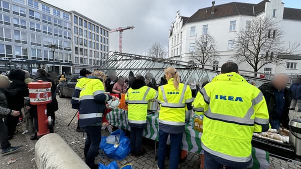 Mehrere IKEA-Mitarbeitende in gelben Warnjacken mit IKEA-Logo verteilen an einem Straßenstand Essen und Getränke an eine Gruppe von Menschen. Die Szene spielt sich in einem städtischen Umfeld mit modernen Gebäuden und einem U-Bahn-Eingang im Hintergrund ab.  