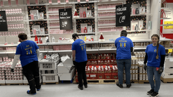 Mehrere Personen der Sozialgruppe Kassel in blauen T-Shirts mit dem Aufdruck „Hej! Wir fördern ein inklusives Miteinander“ sortieren Produkte in einem Regal mit Weihnachtsartikeln. Eine Mitarbeiterin steht lächelnd im Vordergrund, während die anderen sich um die Waren kümmern.  