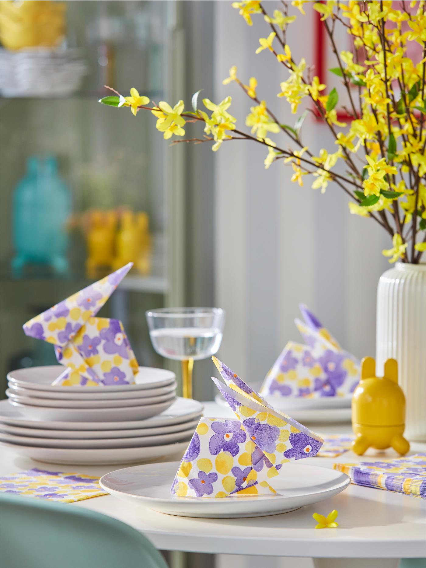 Mehrere weisse Teller stehen auf einem Tisch. Auf den Tellern liegen gefaltete TJÄRLEK Papierservietten mit Blumenmuster.