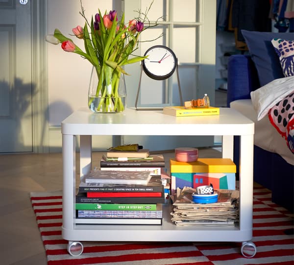 Meja sisi beroda TINGBY berwarna putih dengan banyak buku di bahagian bawah dan vas yang penuh dengan bunga dan jam di bahagian atas.