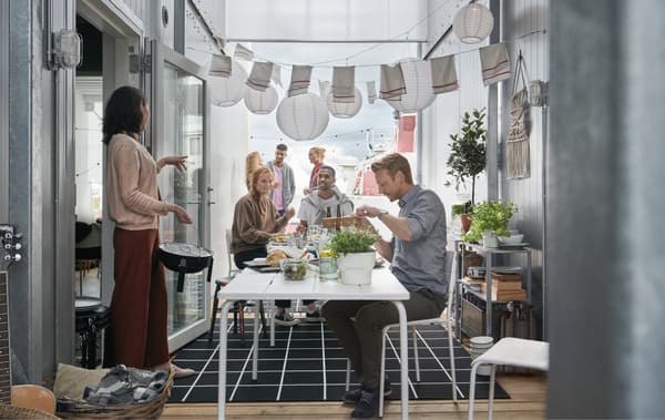 Menschen, die sich rund um VÄDDÖ Tisch für draussen in Weiss versammelt haben. Der Tisch ist gedeckt und steht zwischen zwei Schiffscontainern, darüber hängen Solarleuchten.