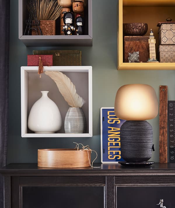 Mesa de apoio com um candeeiro de mesa com coluna Wi-Fi. Arrumação decorativa em diversas estantes de parede pequenas por cima da mesa.