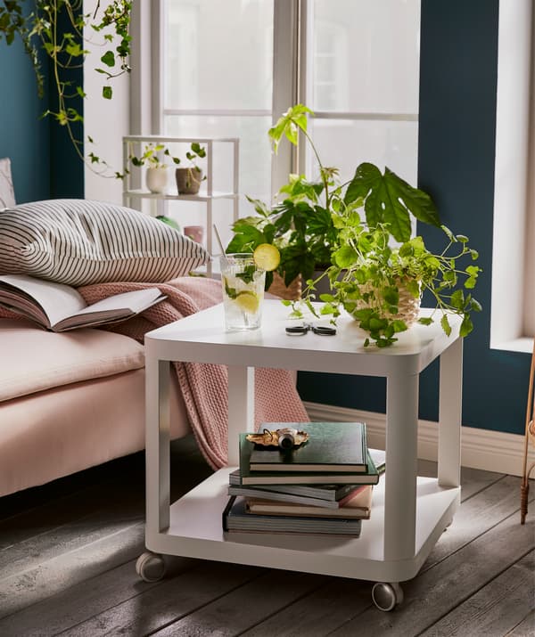 Mesa de apoio em forma de cubo com dois níveis e com rodízios – livros em baixo, plantas e um copo em cima – ao lado de uma chaise longue.