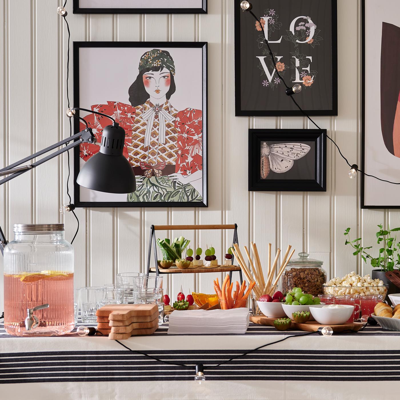 Mesa de bufê com comida e bebidas em várias taças, pratos e recipiente com torneira VARDAGEN, junto a parede com imagens em molduras.