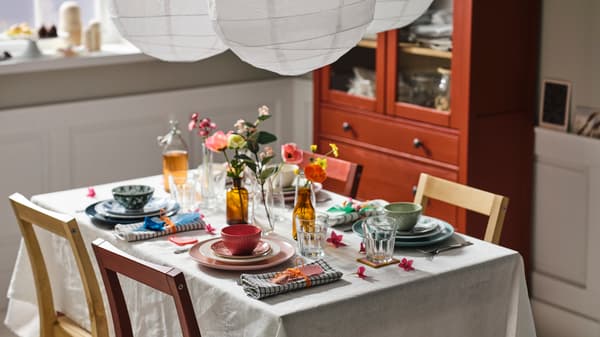 Mesa de comedor con mantel blanco con vajilla de varios colores junto a sillas de madera.
