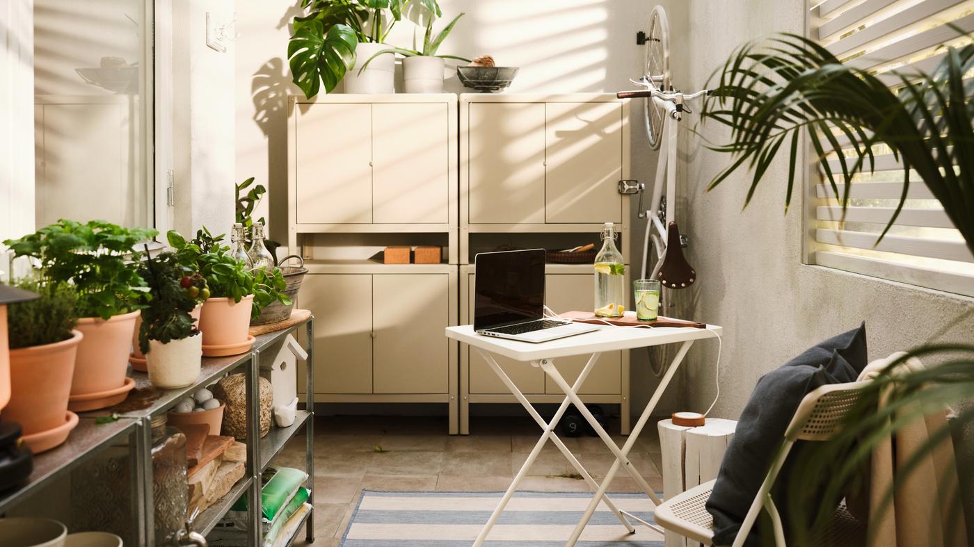 Mesa de refeições para exterior em branco com um computador portátil, quatro armários em bege e em aço empilhados, várias plantas e uma bicicleta em branco pendurada.