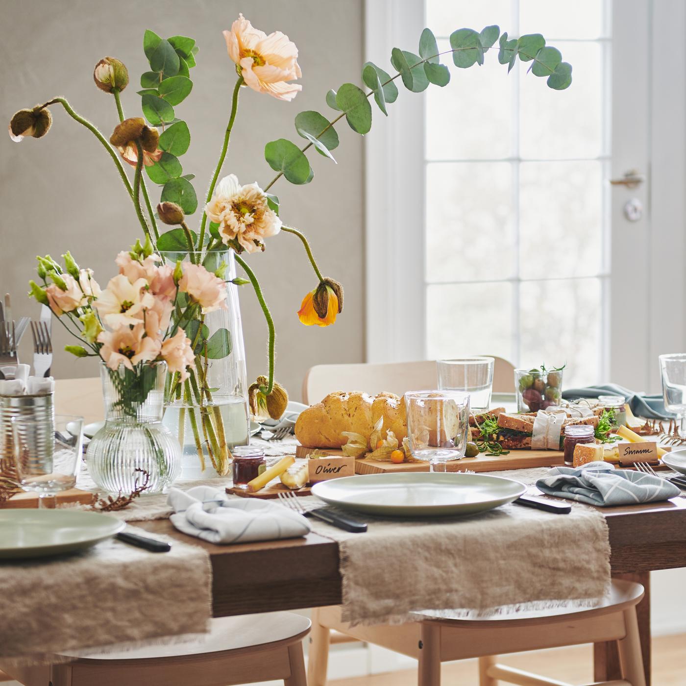 Mesa MÖRBYLÅNGA, em chapa de carvalho, decorada para jantar com loiça FÄRGKLAR em verde mate e dois arranjos de flores em jarras de vidro.