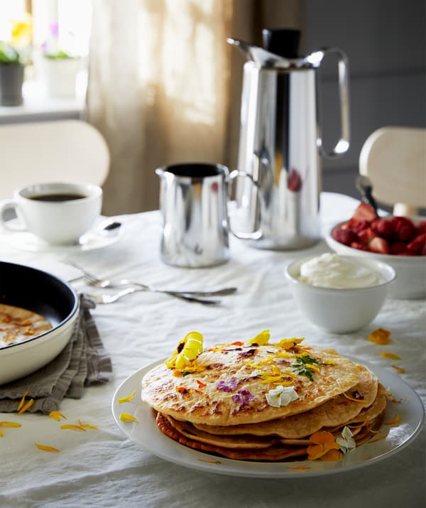 Mesa preparada para pequeno-almoço com toalha, termos com café, morangos com natas e um prato de panquecas guarnecidas com flores.