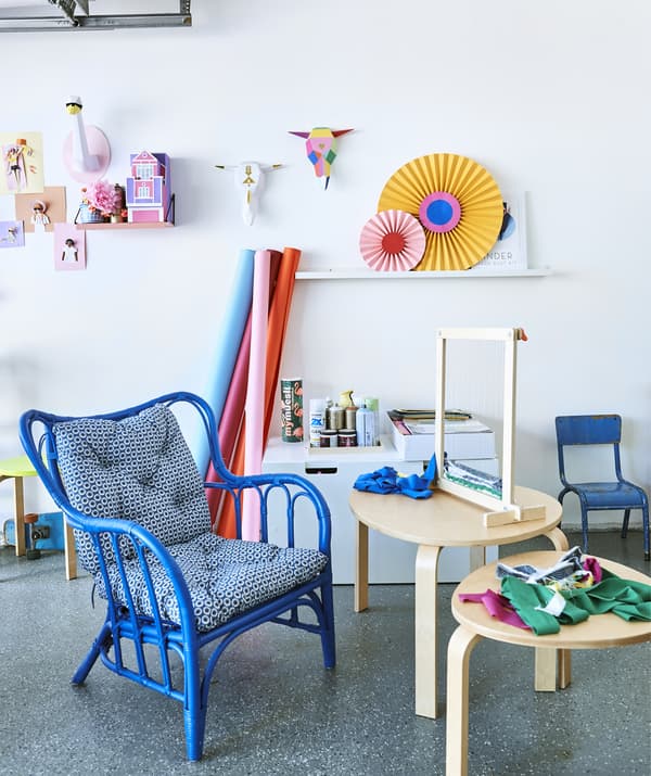 Métier à tisser et tissu sur deux tables d’appoint en bois, fauteuil bleu et œuvres d’art colorées sur un mur blanc.