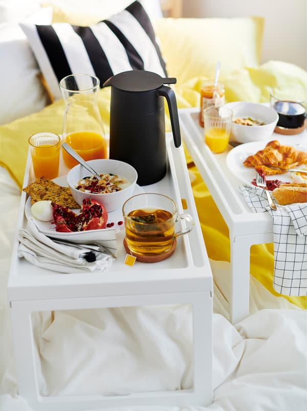 Mic dejun –  suc, croissant, cereale, cafea – servit pe două tăvi de mic dejun KLIPSK așezat pe unui pat ușor deranjat.