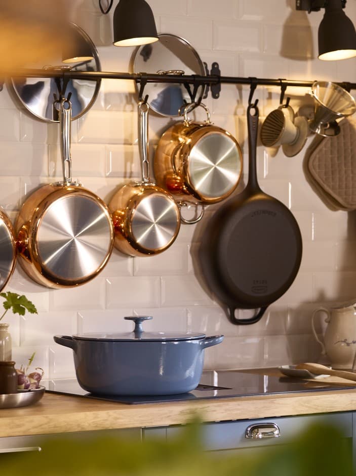 Miedziane patelnie widszące na ścianie i niebieskie naczynie żąroodporne VARDAGEN stojące na szafce kuchennej.