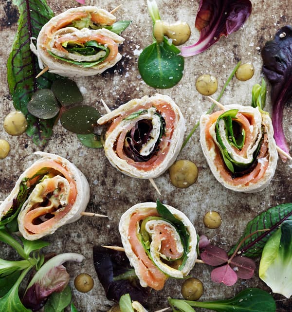 Mini wrap rolls with marinated salmon and mustard sauce served on a marble tray decorated with salad leaves and picked herbs, seen from above.