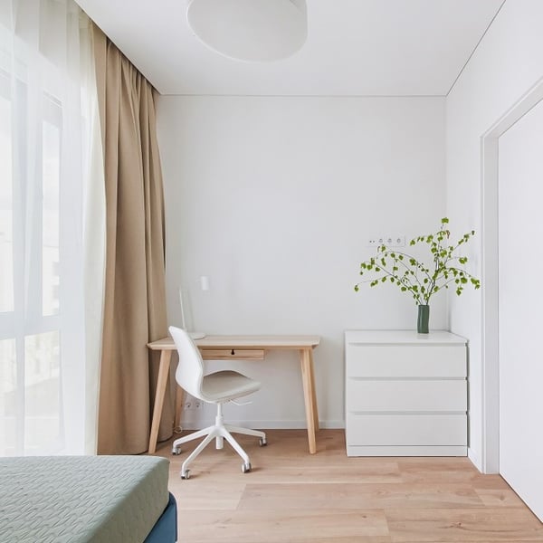 Minimalist home office corner in a bright room with a light wooden floor. A small desk with wooden legs and a white swivel chair sits near beige floor-length curtains and a large window. A modern white chest of drawers with a green vase holding leafy branches is placed next to the desk. The overall color palette is light and neutral, with natural light enhancing the clean, airy feel.