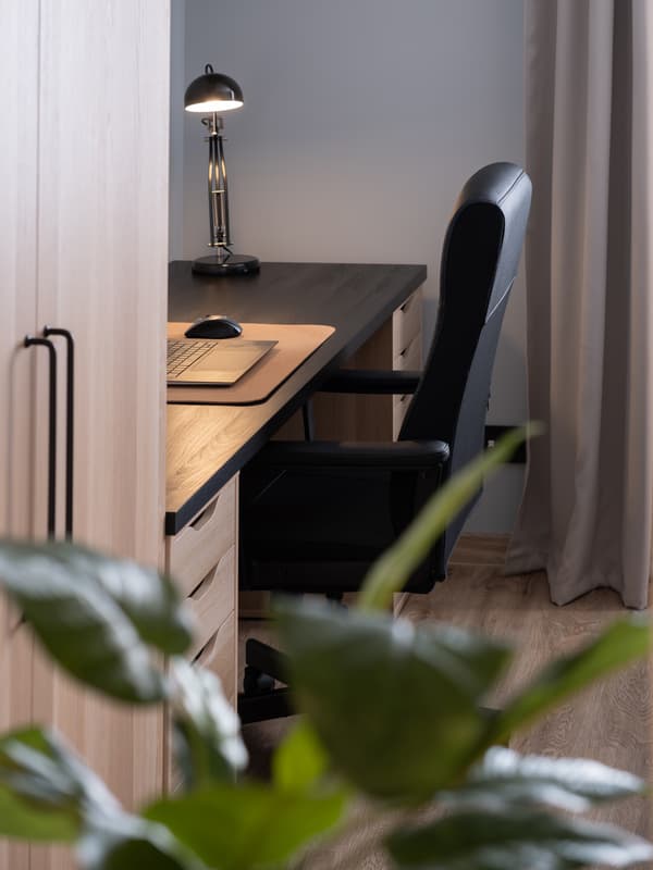 Minimalist home office setup with a wooden desk, black chair, desk lamp, and soft natural light from a nearby window.