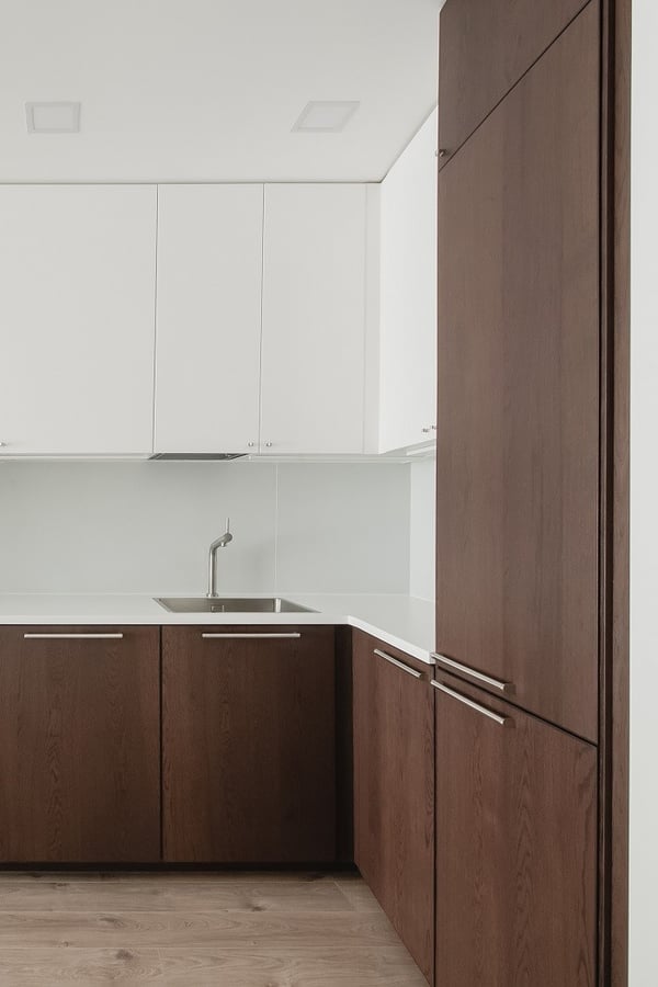 Minimalist kitchen corner featuring white upper cabinets and dark wood lower cabinets with sleek horizontal handles. A modern single-handle faucet is mounted on a white countertop above a stainless steel sink, with a light wood floor enhancing the clean, contemporary design.
