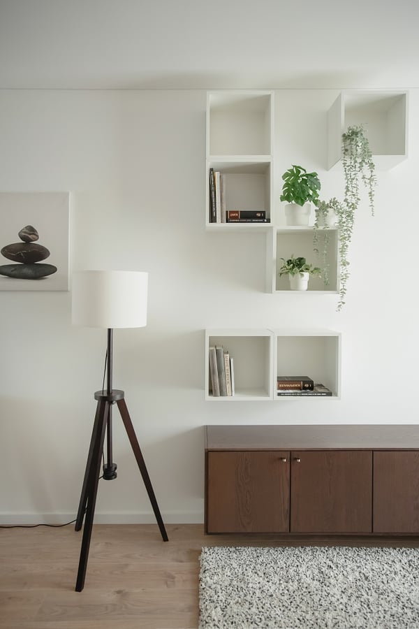 Minimalist wall setup featuring white open cube shelves arranged asymmetrically, displaying books and potted green plants. A dark wooden cabinet sits below, paired with a white lampshade on a tripod black stand. Light wooden flooring and a soft, speckled rug add warmth and texture to the bright interior.