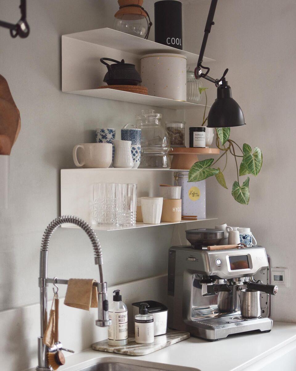 Minimalistische koffiecorner in de keuken met een espressomachine op het aanrecht, witte wandplanken met kopjes en glazen, een zwarte wandlamp, groene plant en een roestvrijstalen kraan.