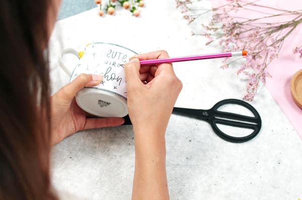 Mit einem Bleistift wird die Vorlage nachgezeichnet sodass das Kohlepapier es auf die Tasse überträgt. 