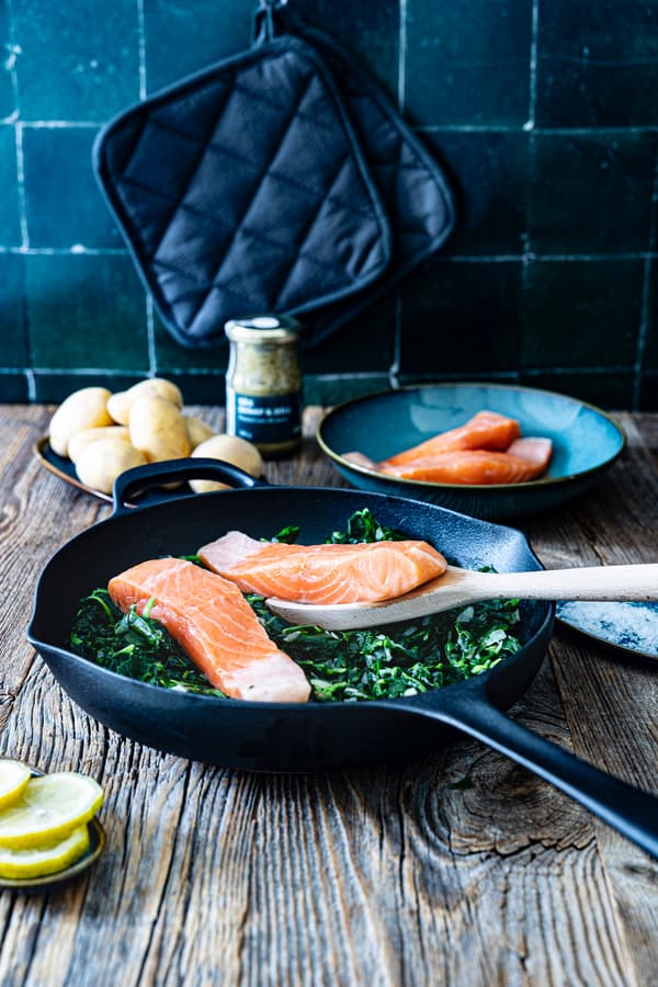 Mit einem Holzlöffel werden zwei Lachsfilets auf den frischen Spinat in der Pfanne gelegt.