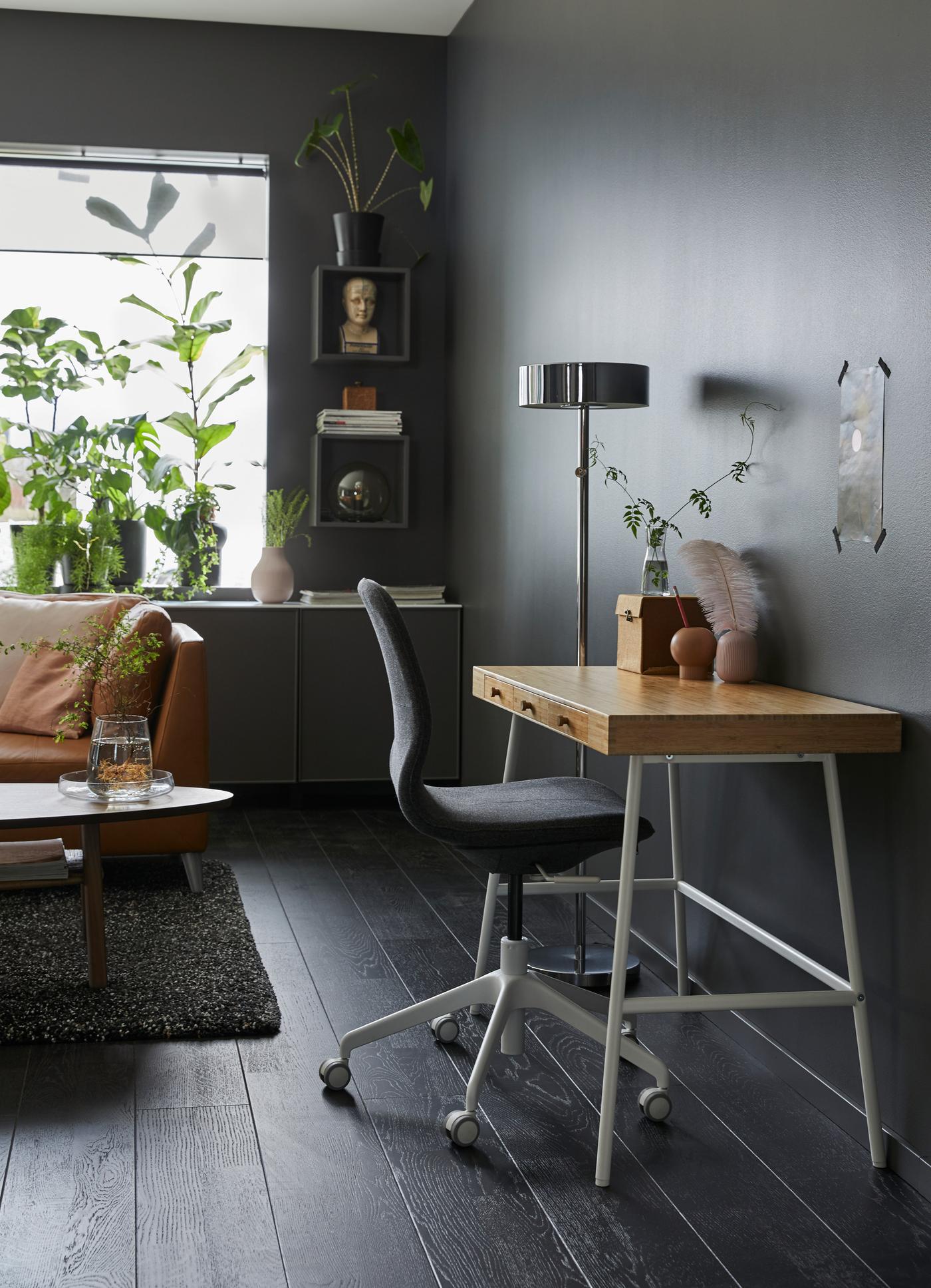 مكتب صغير بسطح خيزران وأرجل بيضاء بمحاذاة حائط غرفة جلوس رمادي داكن.