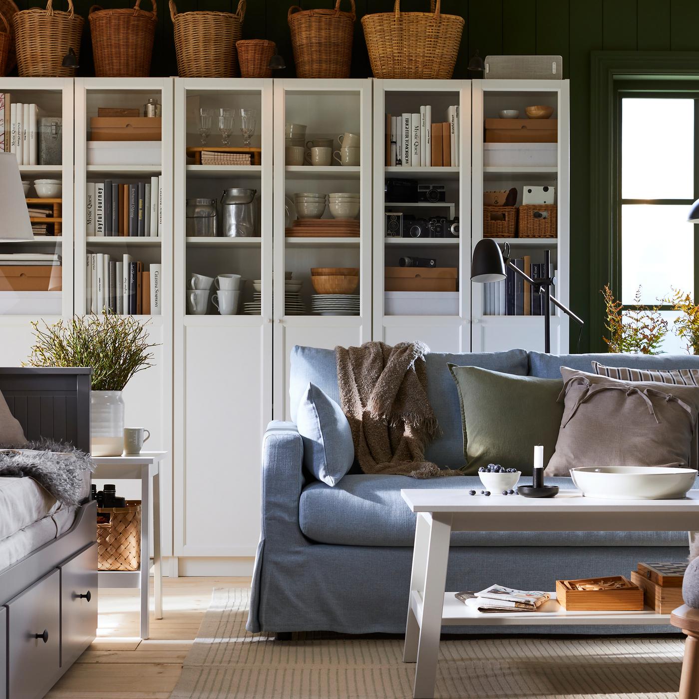 مكتبة BILLY بيضاء بألواح/ أبواب زجاجية مليئة بالكتب والأكواب والأطباق خلف كنبة زرقاء وطاولة قهوة بيضاء.