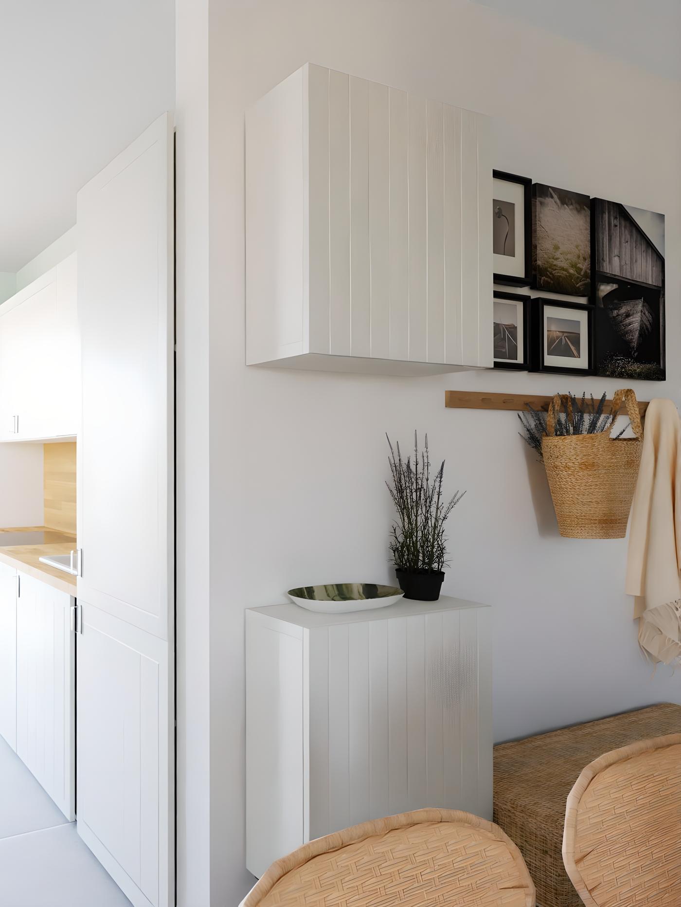 Moderan interijer s bijelim ormarićima, drvenom policom s košarom i kaputom, crno-bijelim fotografijama na zidu, biljkom u crnoj posudi i dekorativnom zdjelom; u pozadini dio kuhinje.