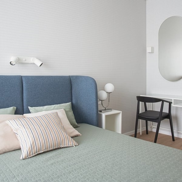 Modern and minimal bedroom featuring a blue upholstered headboard, soft green quilted bedding, and a mix of striped and solid pillows in pastel tones. A white wall-mounted reading light is positioned above the bed, while a small white side table holds a sculptural globe-style lamp. A black chair is placed at a sleek white floating desk, with a large arched mirror on the wall above, adding depth and elegance to the light-toned space.