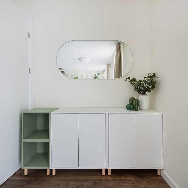 Modern and minimalistic entryway or hallway setup featuring a sleek white cabinet with natural wooden legs. On the left side, an open shelving unit in sage green adds a soft color accent. Above the cabinet, an oval wall mirror reflects a bright room with beige curtains. On the right side of the cabinet, decorative items including a white vase with green foliage and small green ceramic cacti add a fresh and natural touch. The space has a clean, Scandinavian-inspired aesthetic with soft, neutral tones and natural wood flooring.