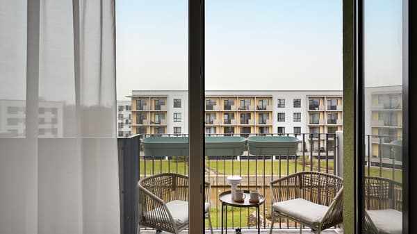 Modern apartment balcony with woven outdoor chairs, a small coffee table, and soft curtains framing a peaceful courtyard view, creating a calm and inviting outdoor living space.