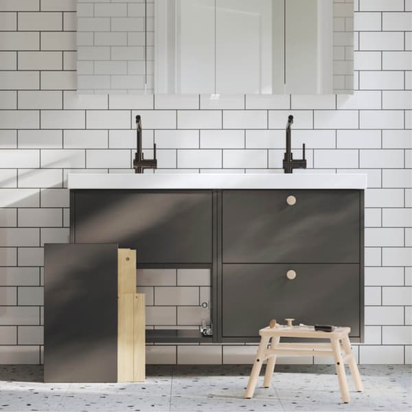 Modern bathroom with double sink, black taps, white wall tiles in brick pattern, dark gray base cabinets with one door removed, and a wooden stool with tools.