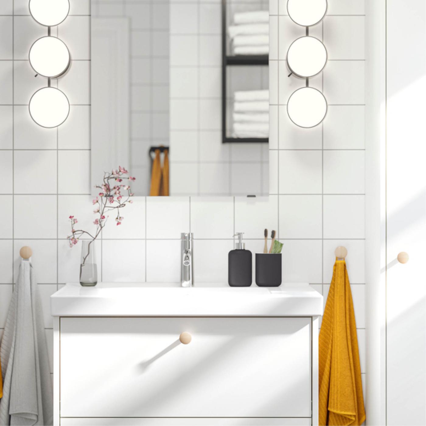 Modern bathroom with white sink, mirror with vertical rows of round lamps, soap dispenser, toothbrush holder, vase with pink flowers and towels on both sides.