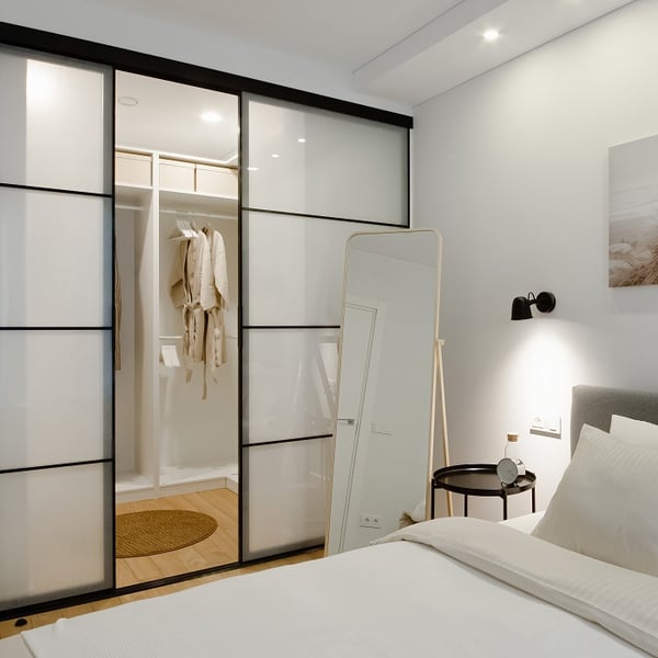 Modern bedroom with a white bed and light bedding, featuring a sliding glass wardrobe with black frames. Inside the wardrobe are hanging beige robes and storage boxes. A full-length mirror stands next to a black bedside table, which holds a clock and carafe under a wall-mounted black sconce.