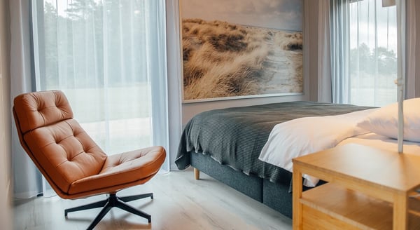 Modern bedroom with leather swivel chair and large nature wall art – Cozy interior featuring a tan leather chair, large framed beach grass artwork, and a neatly made bed with grey bedding.