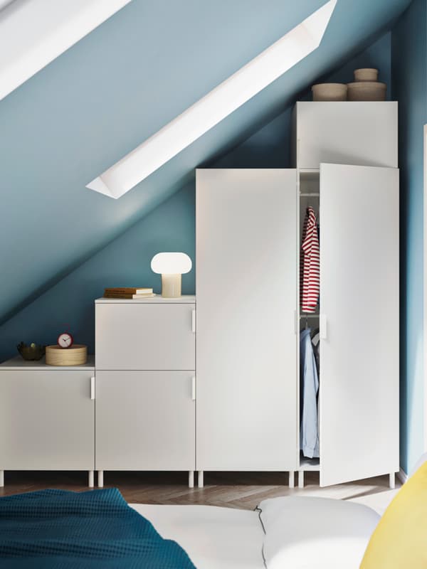 Modern bedroom with slanted ceiling and blue walls, featuring white modular wardrobes, open cabinet with hanging clothes, and a bed with white bedding, blue blanket, and yellow pillow.