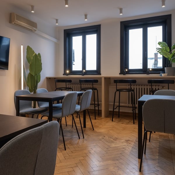 Modern breakroom with natural light coming through two black-framed windows. The space includes dark wood dining tables with light blue upholstered chairs, and a high bar counter with black bar stools along the window wall. A tall green plant, wall-mounted air conditioner, and light herringbone wood flooring enhance the cozy, minimalist interior. Ceiling spotlights provide additional illumination.