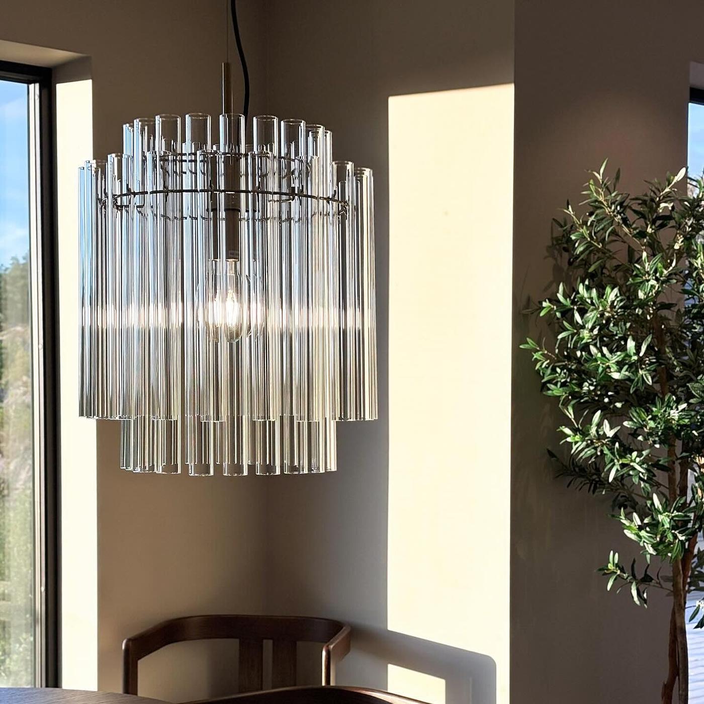 Modern cylindrisk taklampa i glas hänger framför en mörk vägg, med en grön växt och en konsol i bakgrunden.