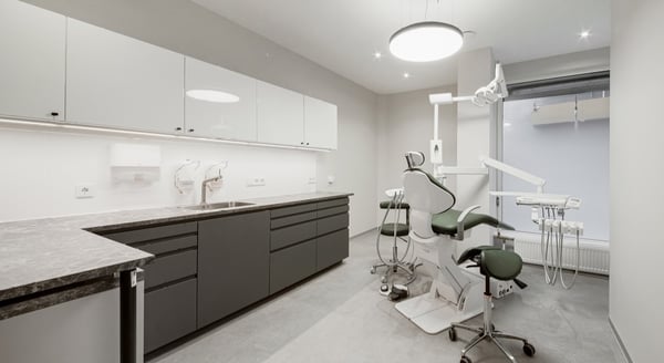 Modern dental treatment room with green dentist chair, sleek cabinets, bright lighting, and large frosted window.