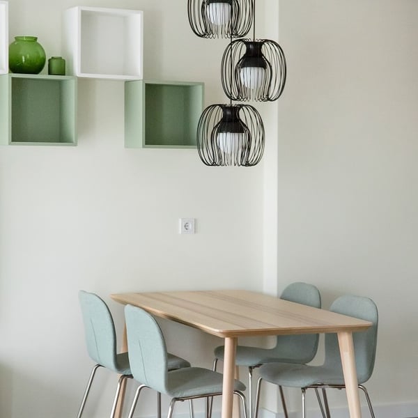 Modern dining area with a light wooden rectangular table surrounded by four mint green upholstered chairs. Above the table hang three decorative black wire pendant lights with white inner bulbs. On the wall behind the table are open cube shelves in light green and white, displaying green decorative vases. The overall color scheme is soft and fresh, with a minimalist Scandinavian design.