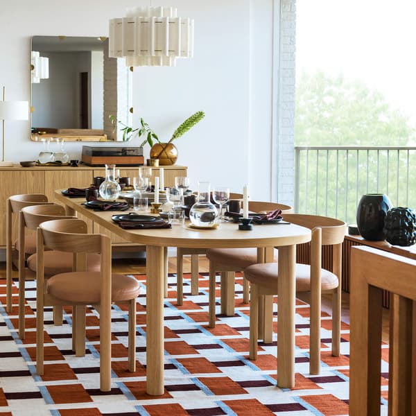 Modern dining room with wooden table set for meal, geometric rug, large window, mirror, and stylish decor on sideboard.