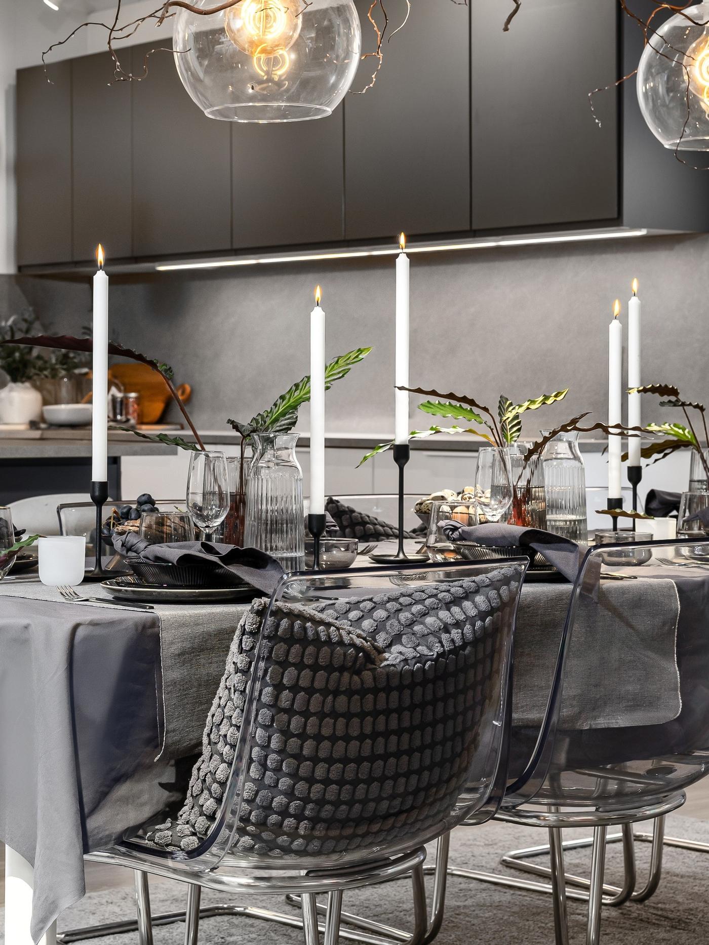 Modern dining table set with a grey tablecloth, black plates, folded linen napkins and tall white candles in minimalist holders.