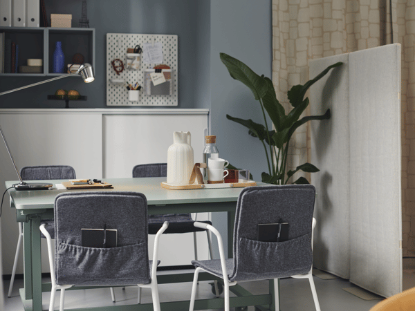 Modern eingerichteter Arbeitsplatz mit IKEA-Tisch, gepolsterten Stühlen mit Taschen an der Rückenlehne, Pflanzen und durchdachter Wandorganisation – ideale Homeoffice-Lösung mit Stil und Funktionalität.