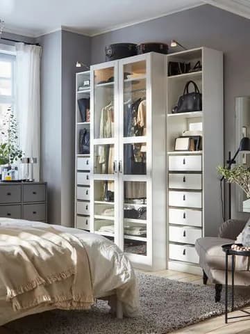 Modern garderob med glasdörrar och lådor fyllda med kläder och väskor. Beige säng och sidobord med blommor i förgrunden.