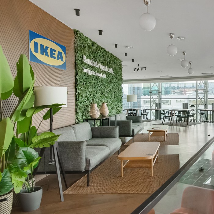 Modern IKEA showroom or office lounge featuring neutral gray sofas, wooden coffee tables, and large green plants. A vertical plant wall with Lithuanian text and the IKEA logo adds a natural focal point. The space is filled with natural light from large windows overlooking a cityscape, and includes additional seating and tables in the background, creating a welcoming and collaborative atmosphere.