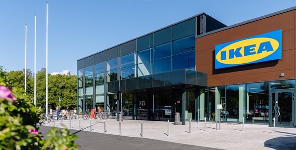 Modern IKEA store exterior with large glass windows and brown facade, featuring a prominent IKEA logo, bicycle racks, and people near the entrance on a sunny day.